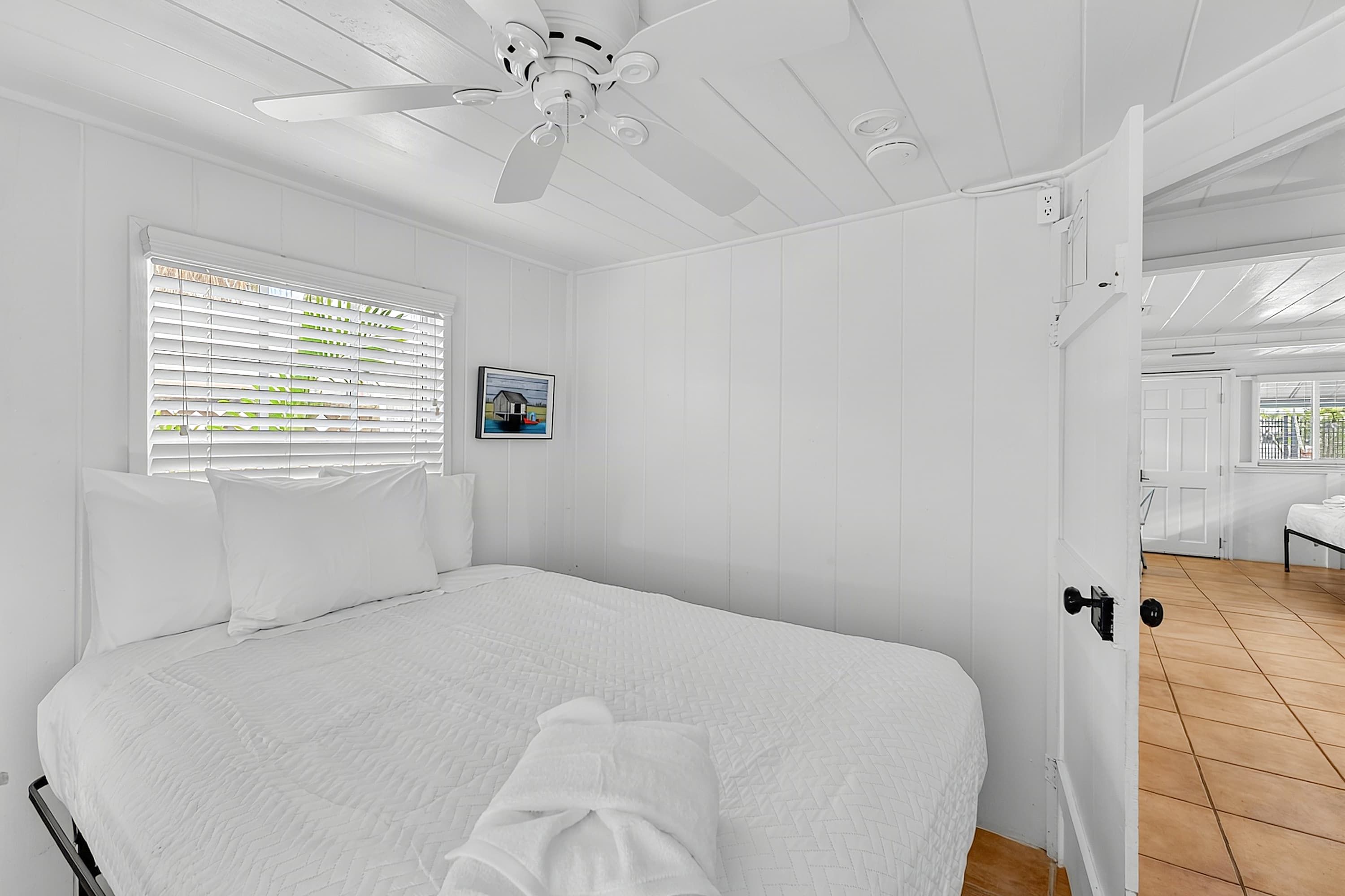Ein Schlafzimmer mit einem Bett, weißen Wänden, einem Deckenventilator, einem Fenster mit Jalousien und einer Tür zu einem anderen Raum.
