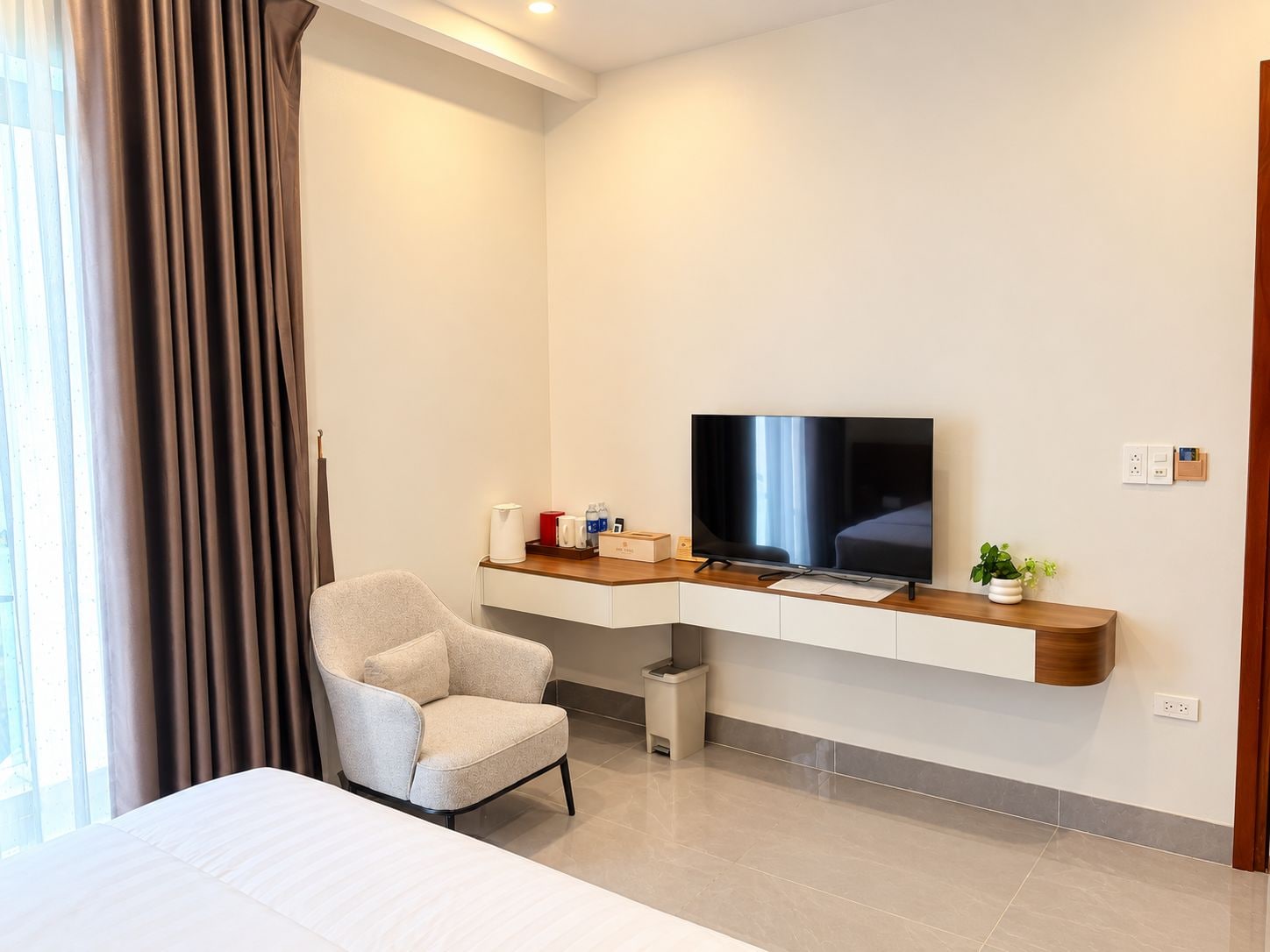 A hotel room with a bed, a TV, a chair, a small table, and a plant.