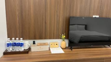Habitación de hotel con cabecera de madera, televisor de pantalla plana, escritorio de madera con bandeja que contiene botellas de agua, taza de café, caja de té y un pequeño jarrón con flores.