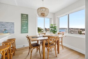 A dining area with a large table and chairs, a hanging light fixture, and a view of the city through large windows. - Beautiful Apartment | Manly Beach Views & Parking (Queenscliff)
