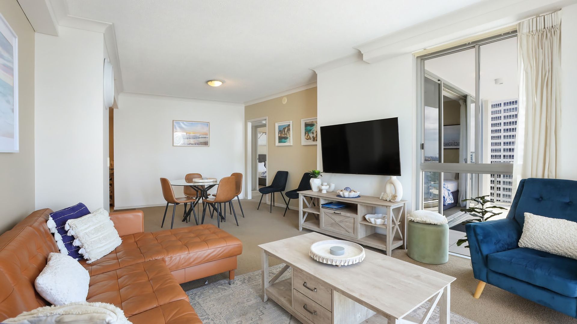 Un salon moderne avec un canapé en cuir, un fauteuil bleu, un coin repas avec des chaises, un téléviseur à écran plat et une grande fenêtre avec des rideaux.