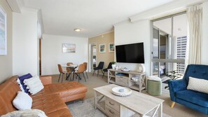 A modern living room with a leather sofa, a blue armchair, a dining area with chairs, a flat-screen TV, and a large window with curtains. - Chevron Renaissance Apartment 2172 (Surfers Paradise)