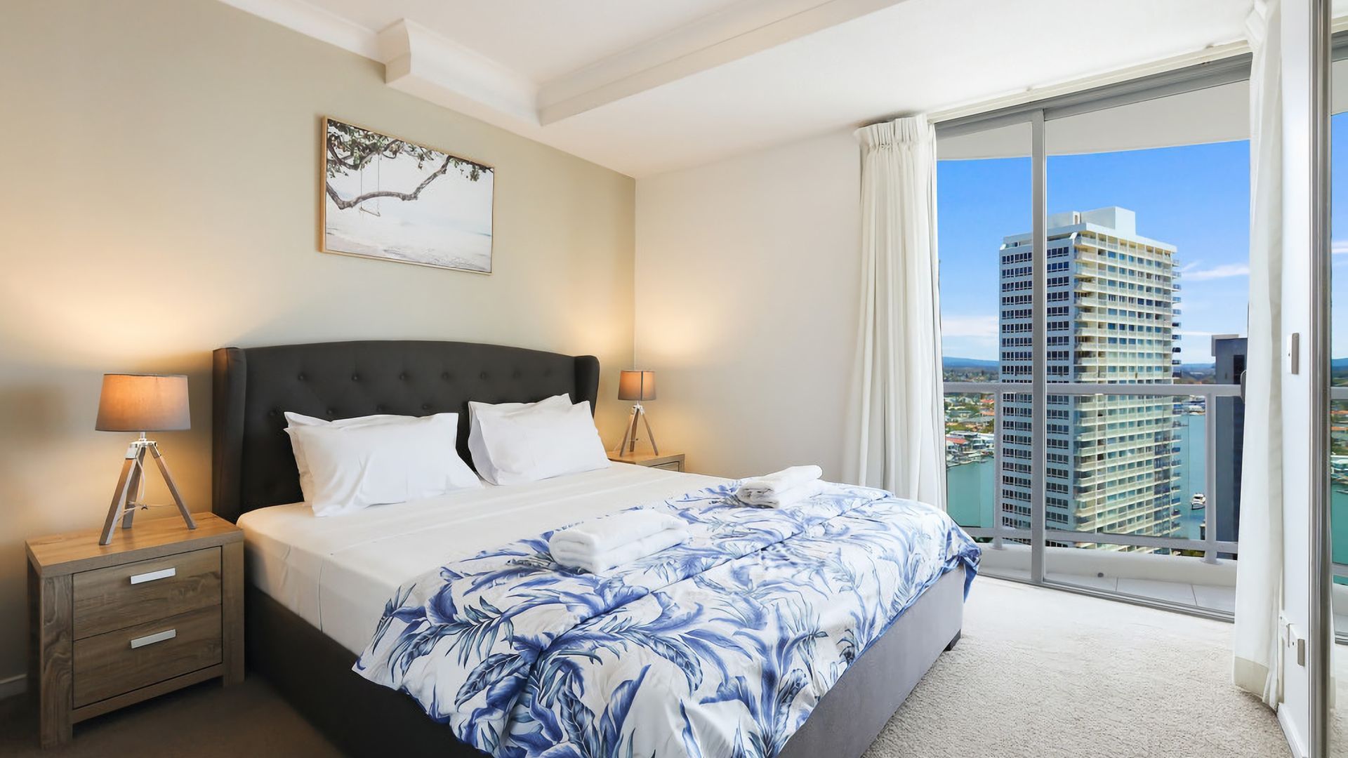 A hotel room with a large bed, two bedside lamps, a nightstand, a window with a view of a cityscape, and a painting on the wall.