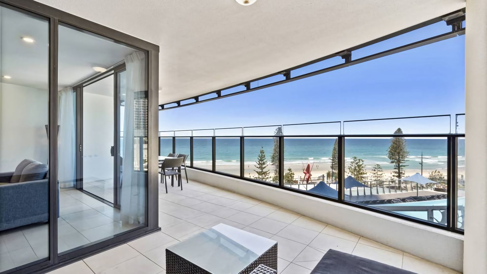 A balcony with a glass door leading to a room, a table, and chairs, overlooking a beach with umbrellas and trees.