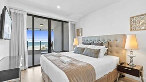 A hotel room with a large bed, a nightstand, a lamp, a window with a view of the beach, and a sliding door. - Esplanade Soul Apartment 402 (Surfers Paradise)