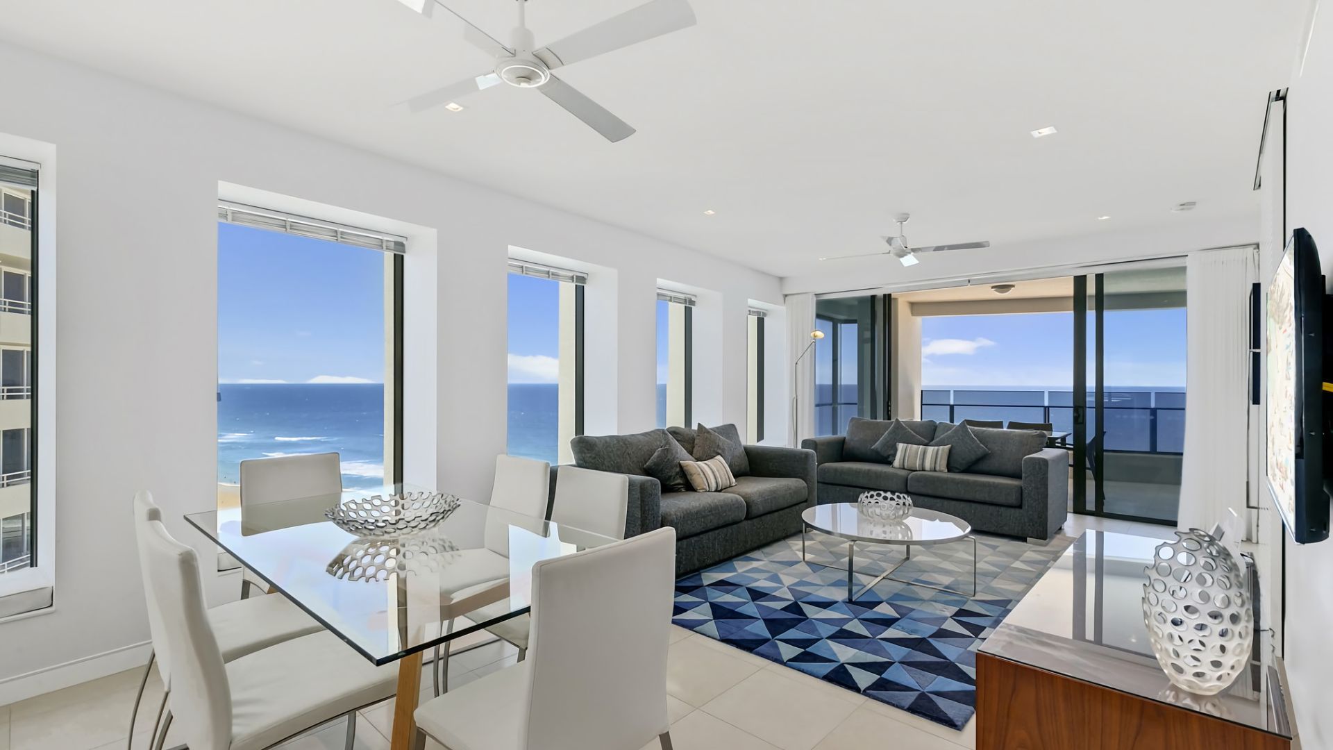 A modern living room with a glass dining table, two sofas, and a view of the ocean.