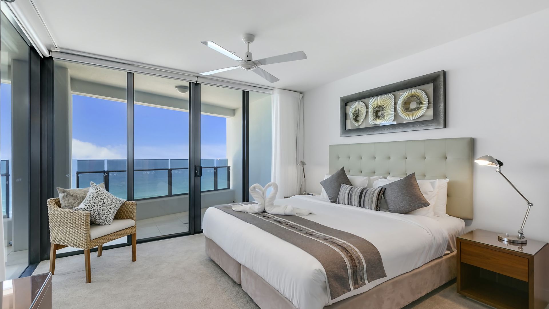 A hotel room with a bed, a chair, a nightstand, a ceiling fan, and a large window with a view of the ocean.