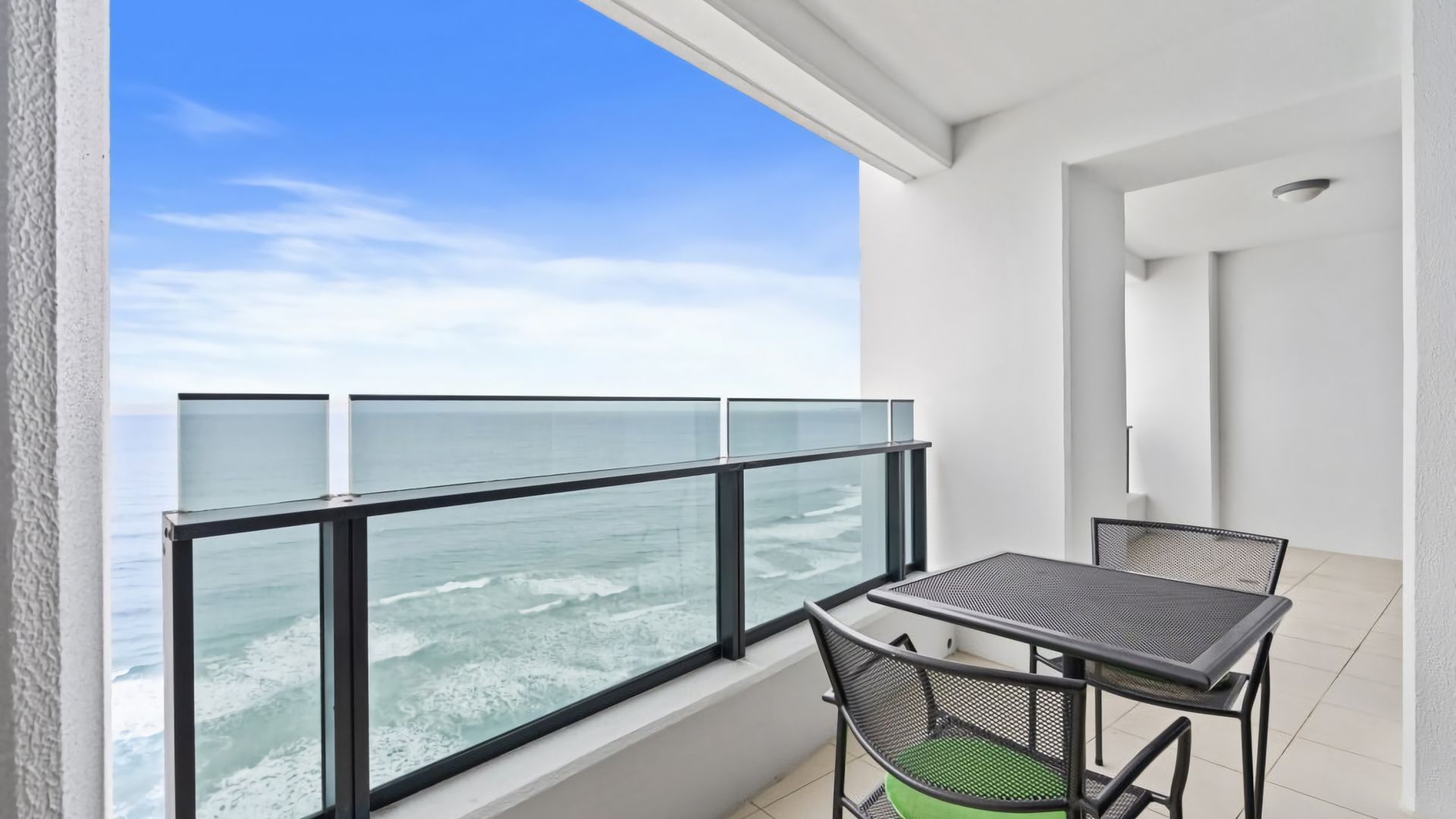A balcony with a glass railing overlooking the ocean, a small table, and two chairs.