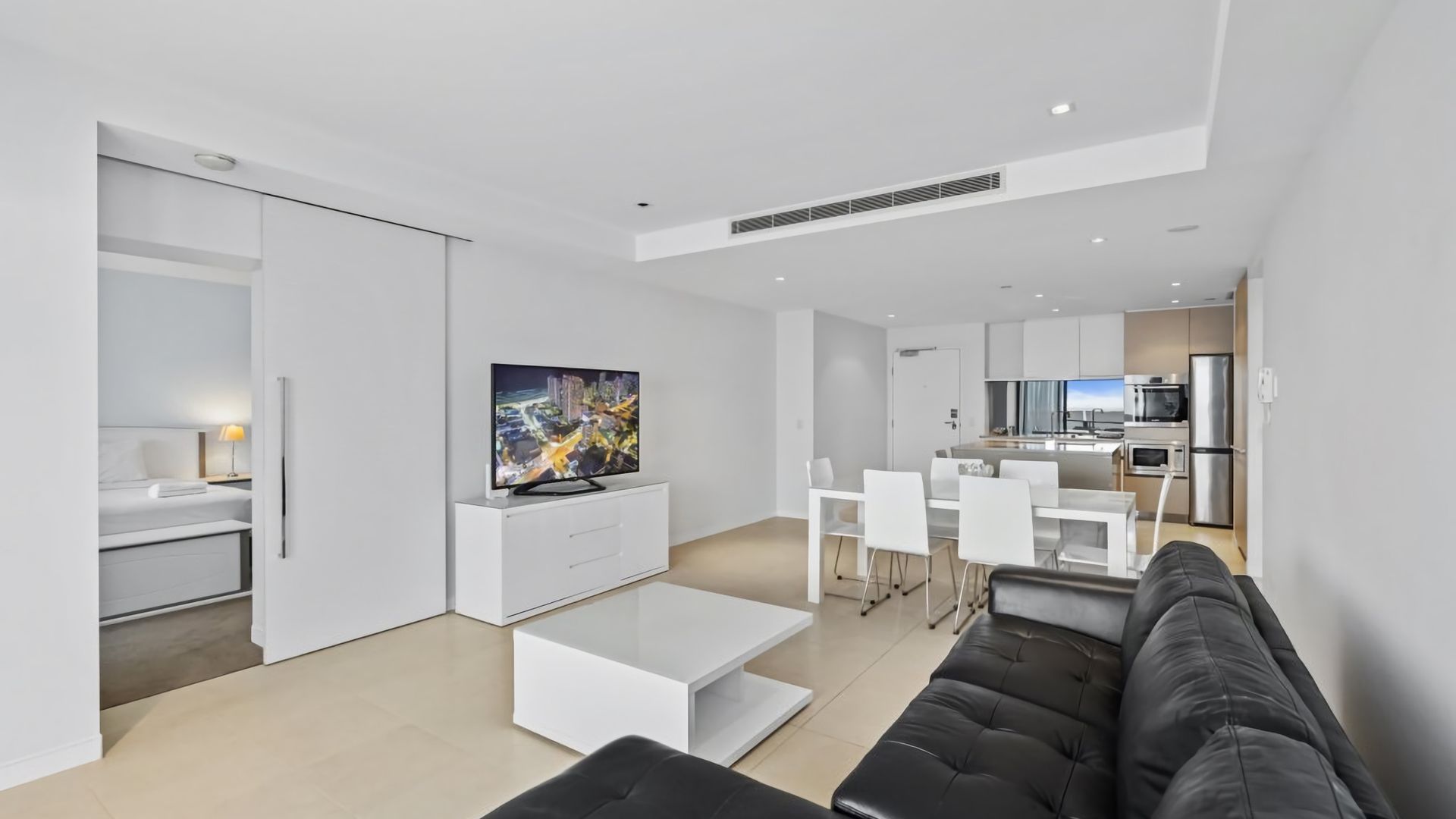 A modern living room with a flat-screen TV, a sofa, and a dining area with white chairs.