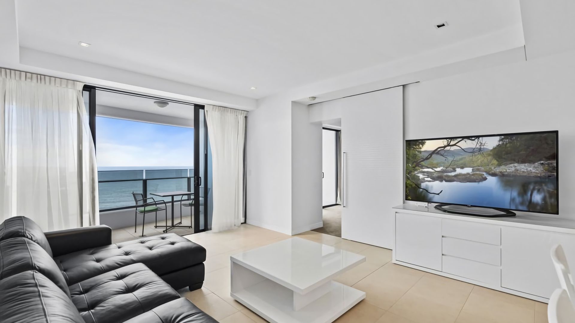 A modern living room with a large flat-screen TV, a black leather sofa, a glass coffee table, and a balcony with a view of the sea.
