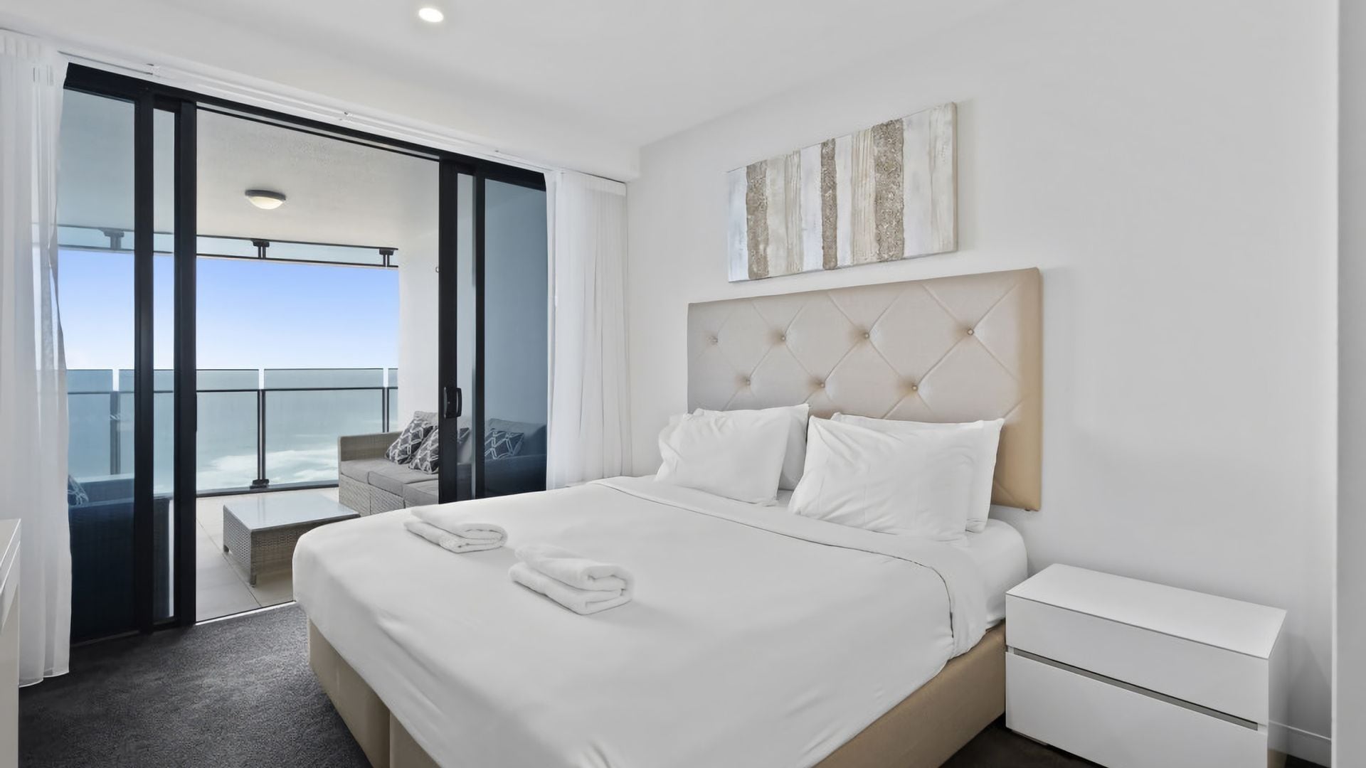A modern hotel room with a large bed, a bedside table, a view of the balcony, and a piece of abstract art on the wall.