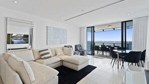 A modern living room with a sectional sofa, a dining area with chairs, and a large window offering a view. - Esplanade Soul Apartment 2502 (Surfers Paradise)