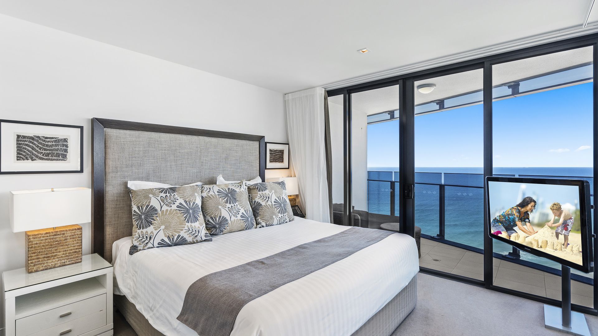 Een moderne hotelkamer met een groot bed, een nachtkastje, een bijzettafeltje, een groot raam met uitzicht op zee en een televisiescherm waarop een zandstrand te zien is.