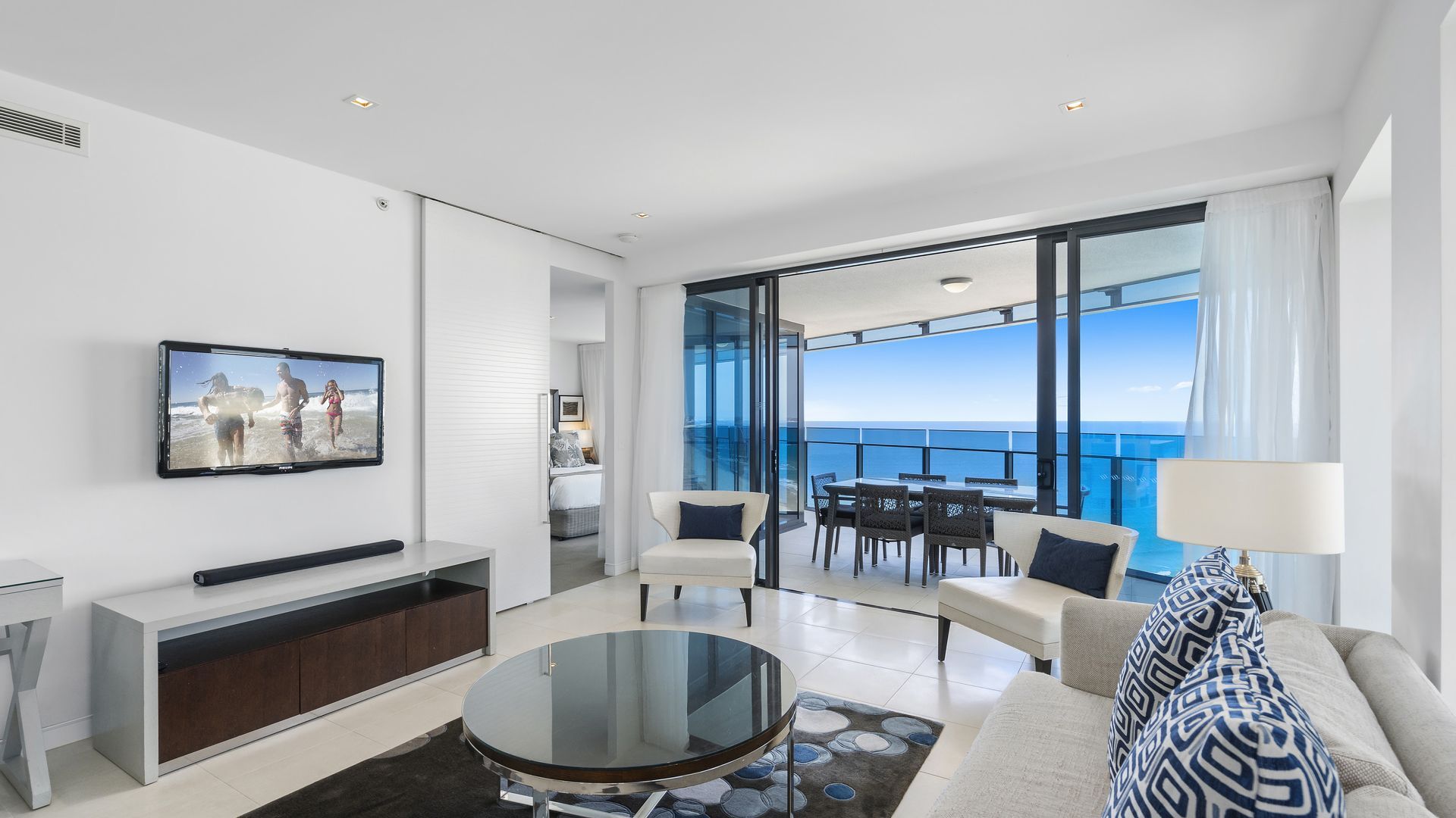 A modern living room with a large TV, a round glass coffee table, and a view of the ocean through large windows.