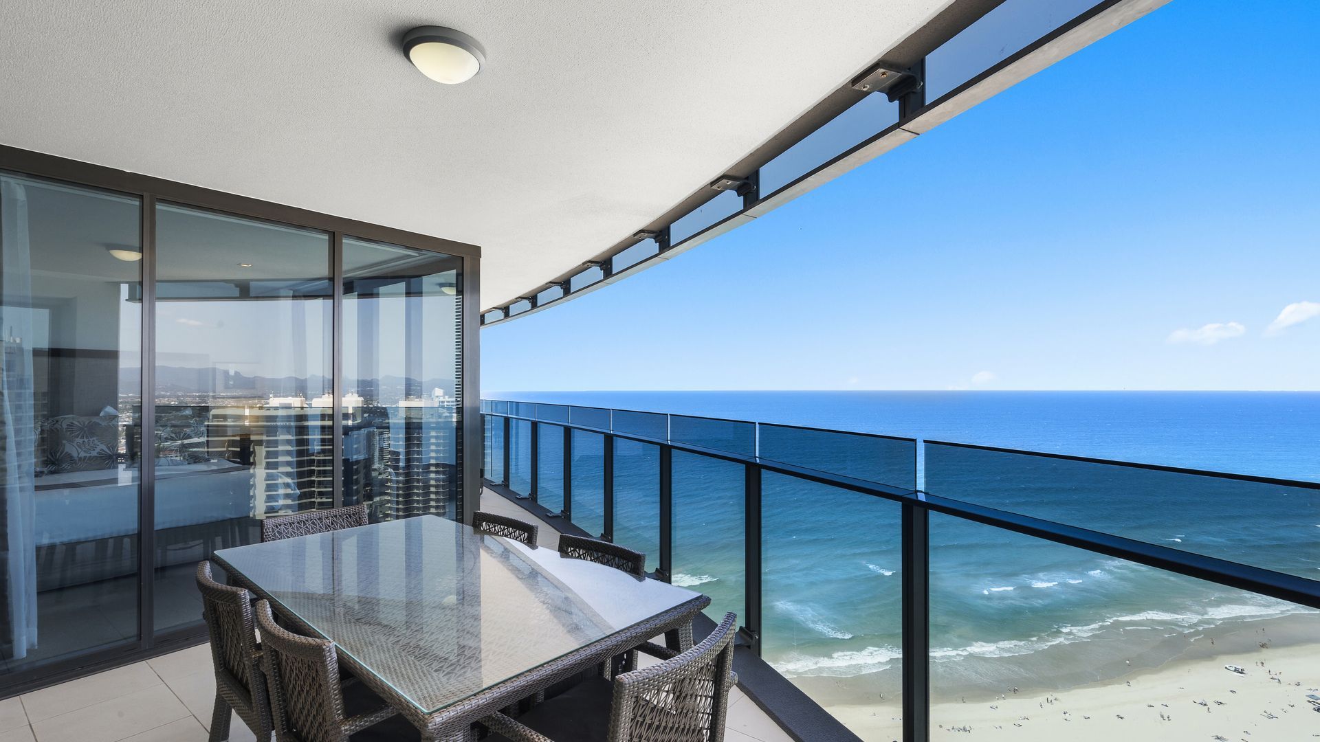 A balcony with a glass table and chairs overlooking a beach and ocean.