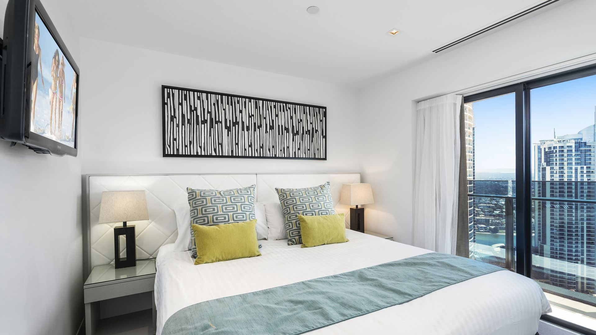 A hotel room with a large bed, two bedside tables with lamps, a city view, and a framed abstract artwork on the wall.