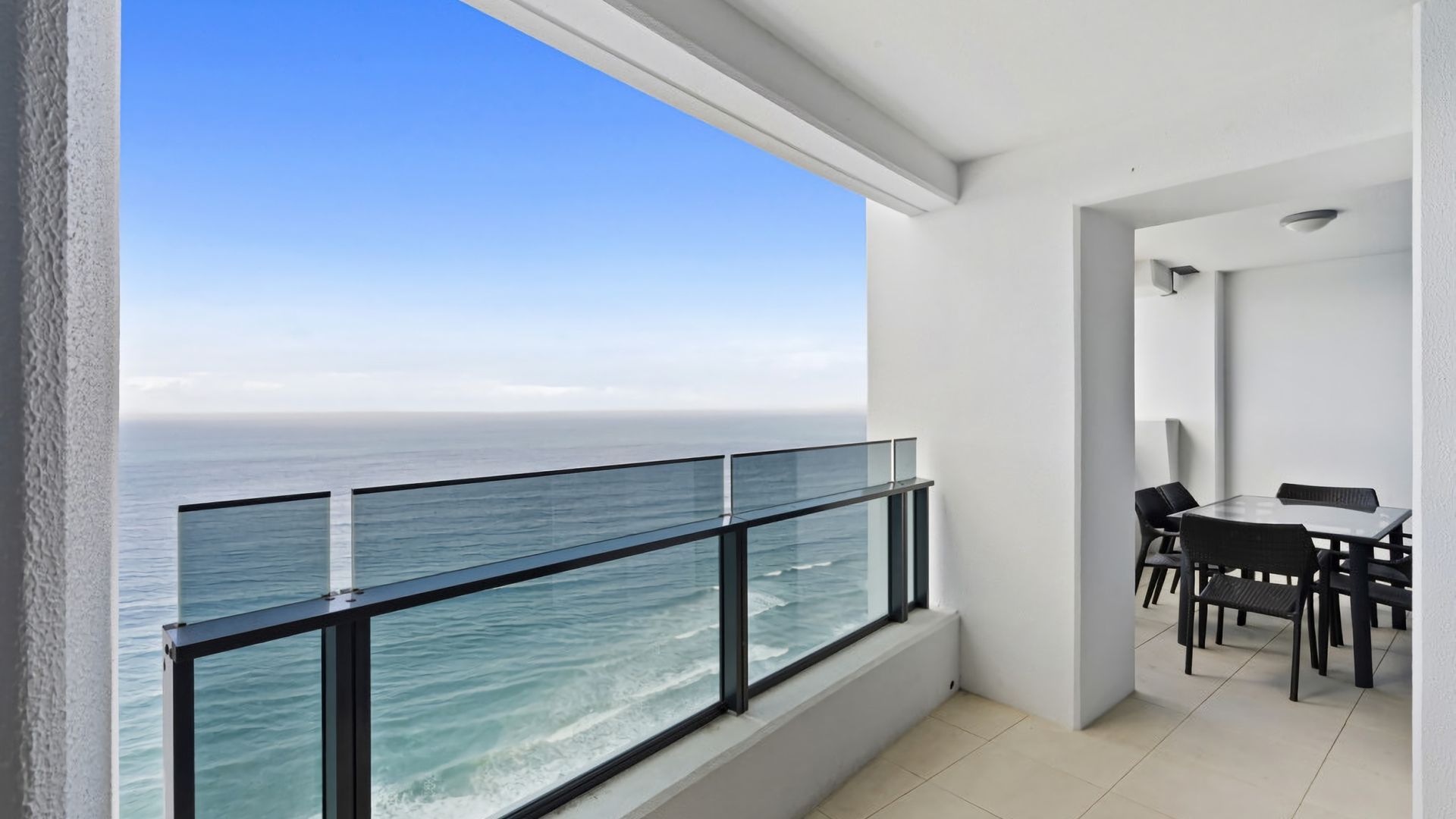 A balcony with a glass railing overlooking the ocean, a dining area with a table and chairs, and a white interior.