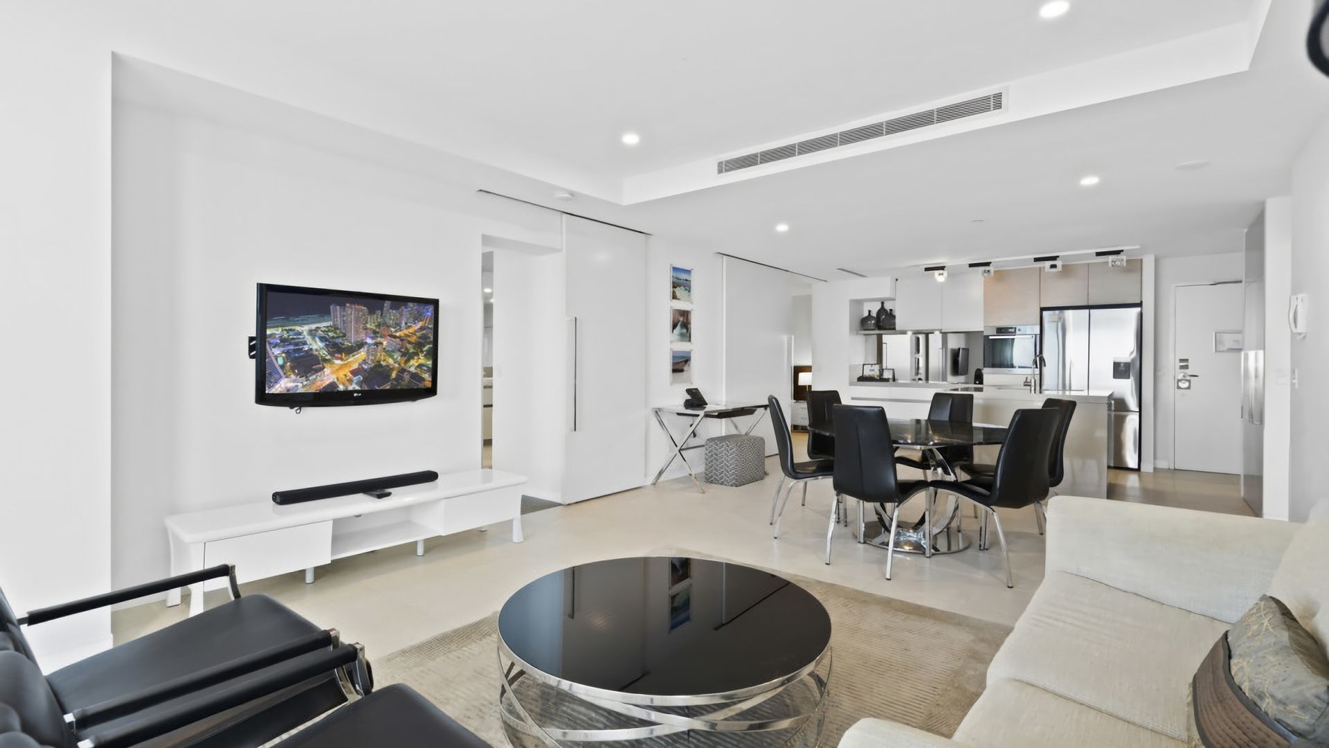 A modern living room with a flat-screen TV, a dining area with a table and chairs, and a kitchen in the background.
