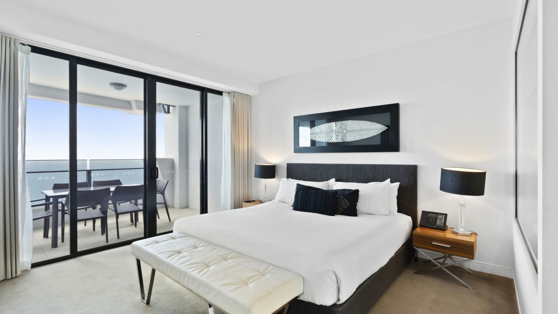 A modern hotel room with a large bed, a balcony with a dining area, and a view of the sky.