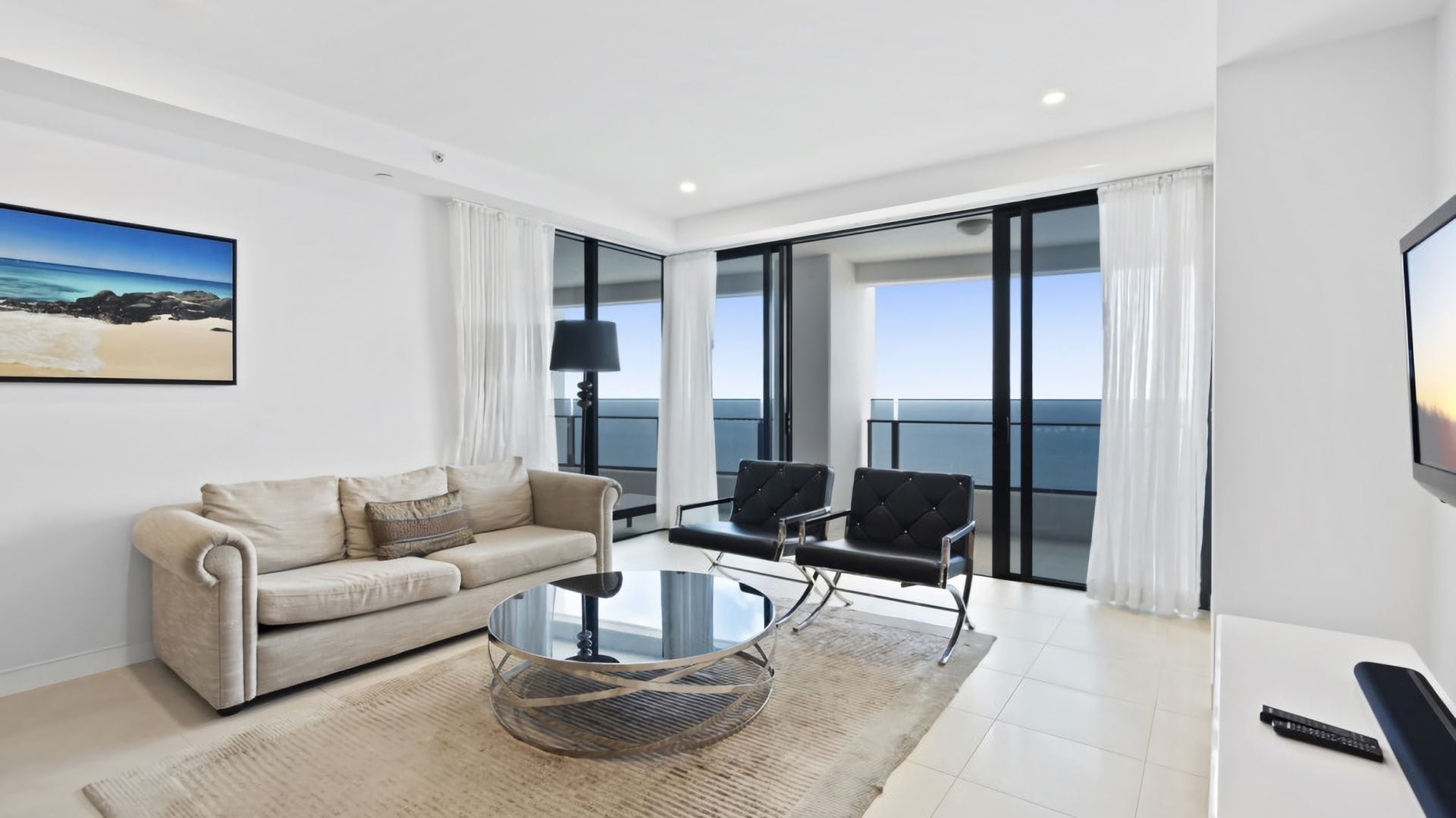 A modern living room with a sofa, a glass coffee table, and a TV. There is a balcony with a view of the sea.