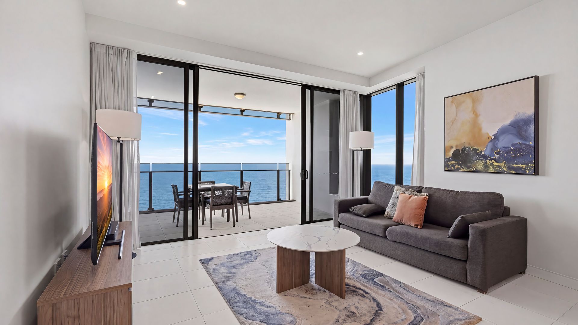 A modern living room with a balcony overlooking the sea, a flat-screen TV, a sofa, and a coffee table.