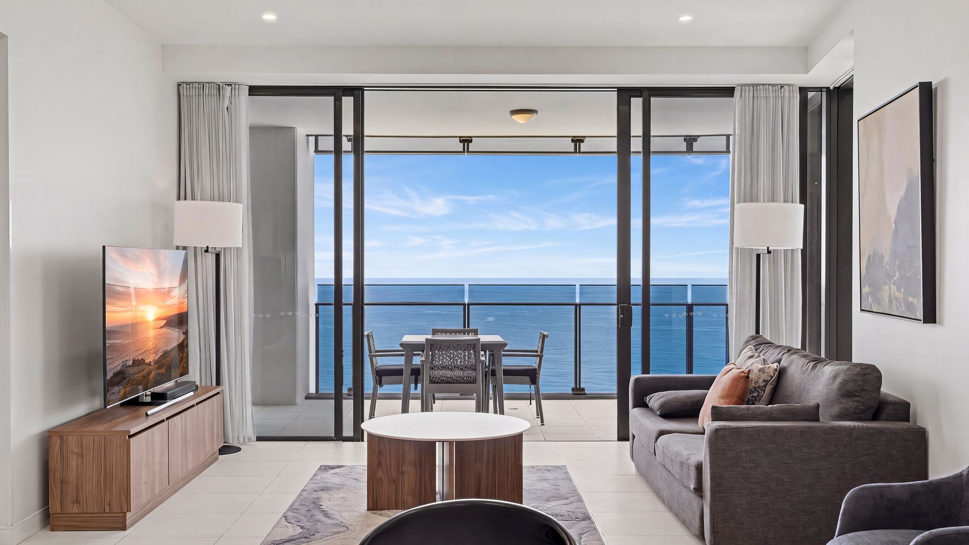 A modern living room with a balcony overlooking the sea, furnished with a sofa, a table, and a TV.