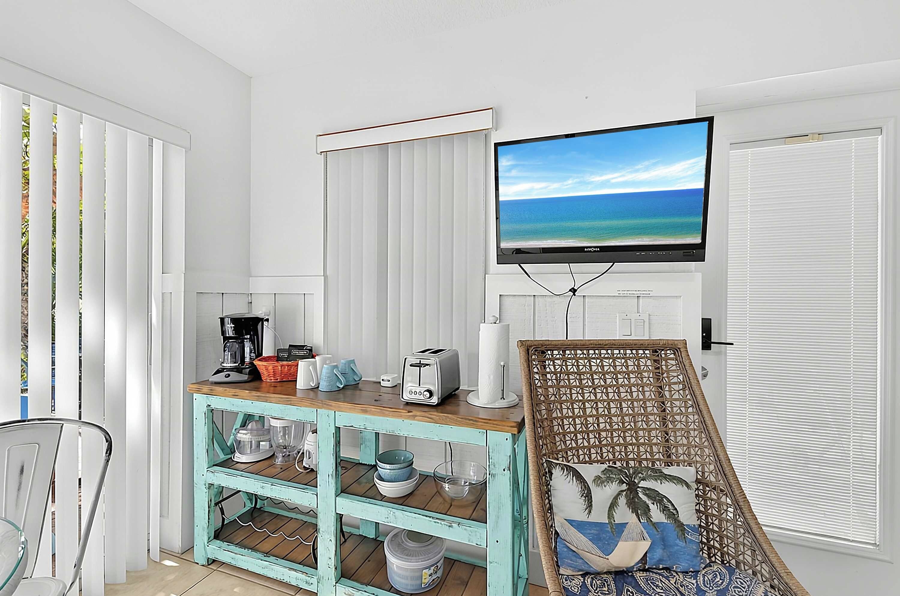 Une pièce avec un téléviseur fixé au mur, diffusant une scène de plage, une machine à café, un grille-pain et un fauteuil en osier agrémenté d’un coussin sur le thème de la plage.