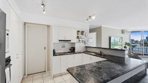 A modern kitchen with white cabinets, a large island, and a city view through the window. - Moorings Apartment 39 (Surfers Paradise)