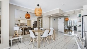 A modern kitchen with white cabinets, a dining area with a wooden table and white chairs, and a large window providing a view of the city. - Moorings Apartment 39 (Surfers Paradise)