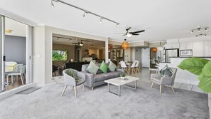 A modern living room with a grey sofa, coffee table, and chairs, a dining area with a table and chairs, and a kitchen with white cabinets. - Moorings Apartment 39 (Surfers Paradise)