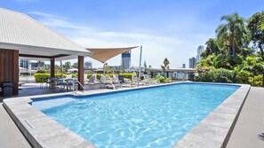 A rooftop swimming pool with lounge chairs and a covered seating area, surrounded by greenery and city skyline views. - Moorings Apartment 39 (Surfers Paradise)