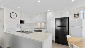 A modern kitchen with white cabinetry, a stainless steel refrigerator, and a wooden dining table. - Moroccan Apartment 143 (Surfers Paradise)