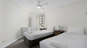 A hotel room with two beds, a ceiling fan, a window with a view of buildings, a bedside table with a lamp, and a wardrobe. - Moroccan Apartment 143 (Surfers Paradise)