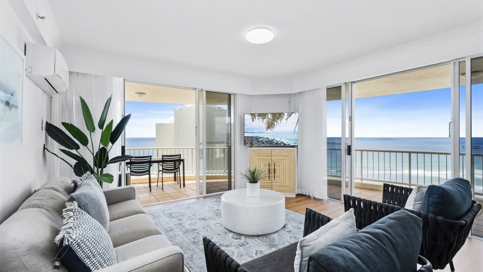 A modern living room with a sofa, chairs, a coffee table, and a TV. There is a balcony with a view of the sea.