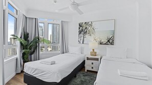 Ein Hotelzimmer mit zwei Betten, einem Deckenventilator, einem Fenster mit Vorhängen, einer Pflanze und einem Bild an der Wand.