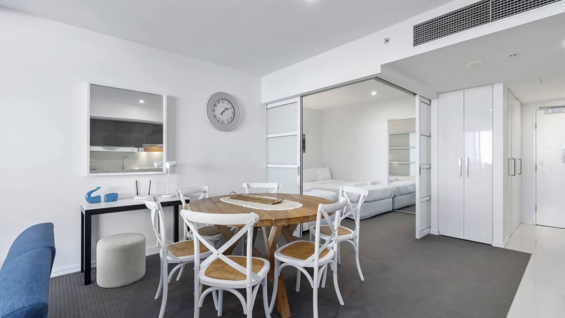 A modern, well-lit room with a round wooden table surrounded by white chairs, a large mirror, and a clock on the wall.