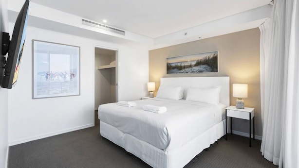 Une chambre d’hôtel avec un lit, deux lampes de chevet, un téléviseur fixé au mur, un tableau au-dessus du lit et une porte donnant sur un placard.