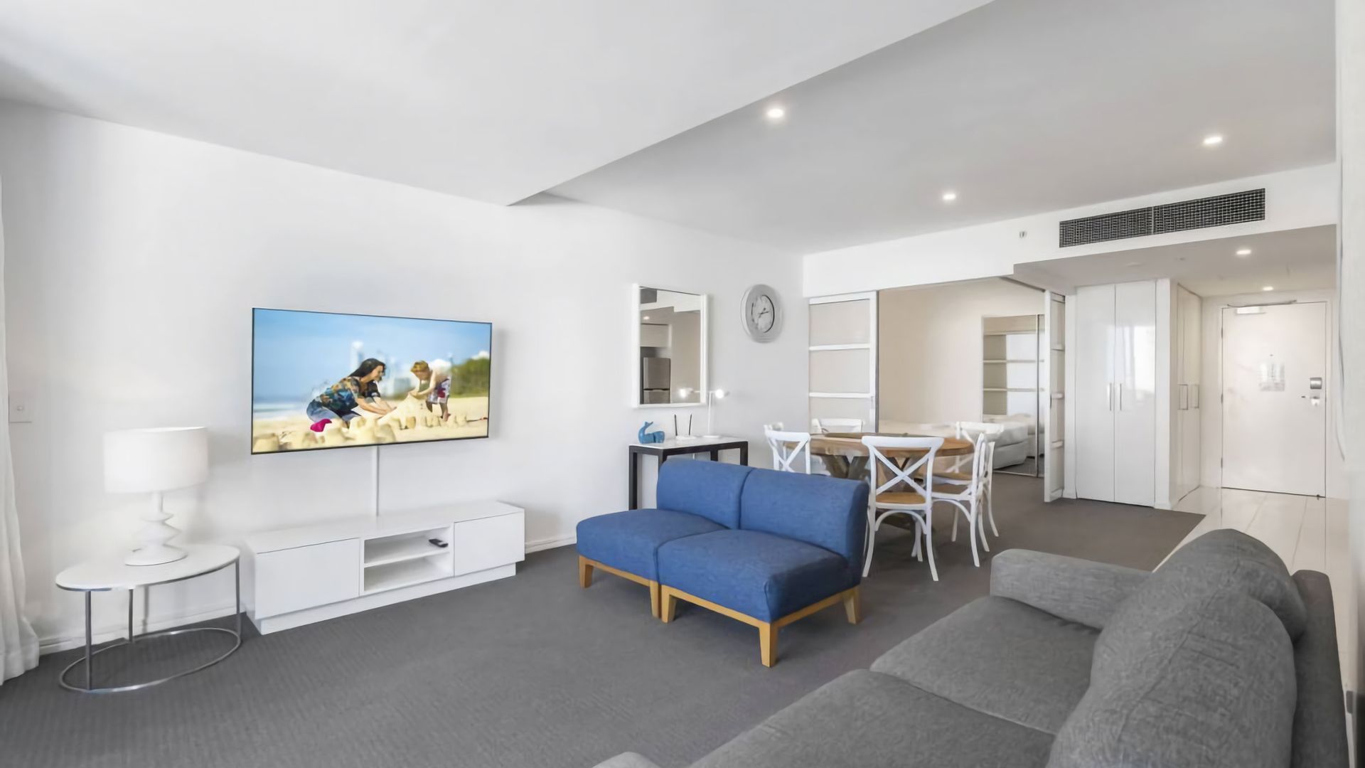 A modern living room with a flat-screen TV, a blue sofa, a grey sofa, a dining table with chairs, and a white cabinet.