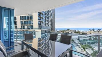 A balcony with a glass table and chairs, overlooking a cityscape and the ocean.