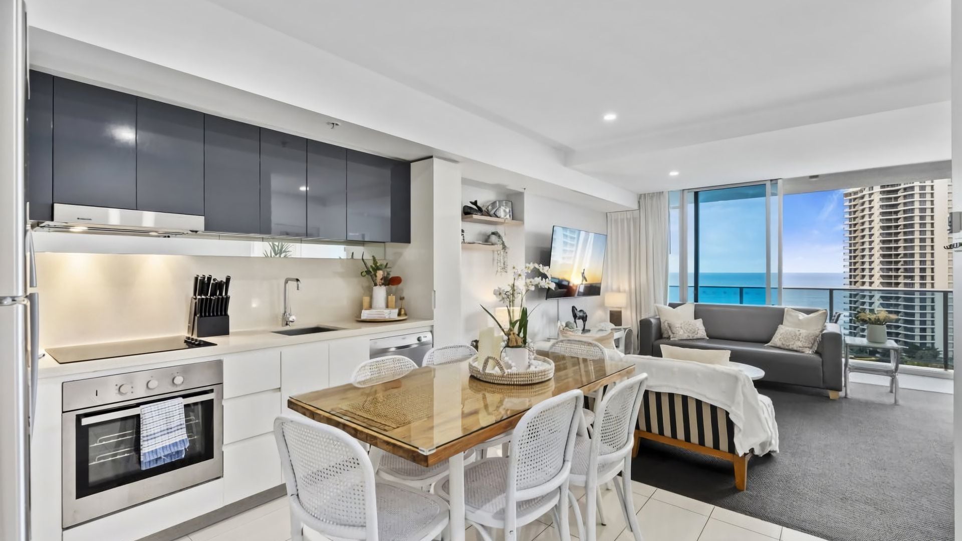 A modern kitchen with white cabinets, a dining area with a wooden table and white chairs, and a living room with a grey sofa and a view of the ocean.