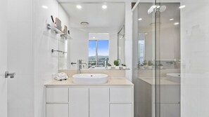A modern bathroom with a large mirror, a white sink, and a glass shower enclosure. - Orchid Residences Apartment 11405 (Surfers Paradise)