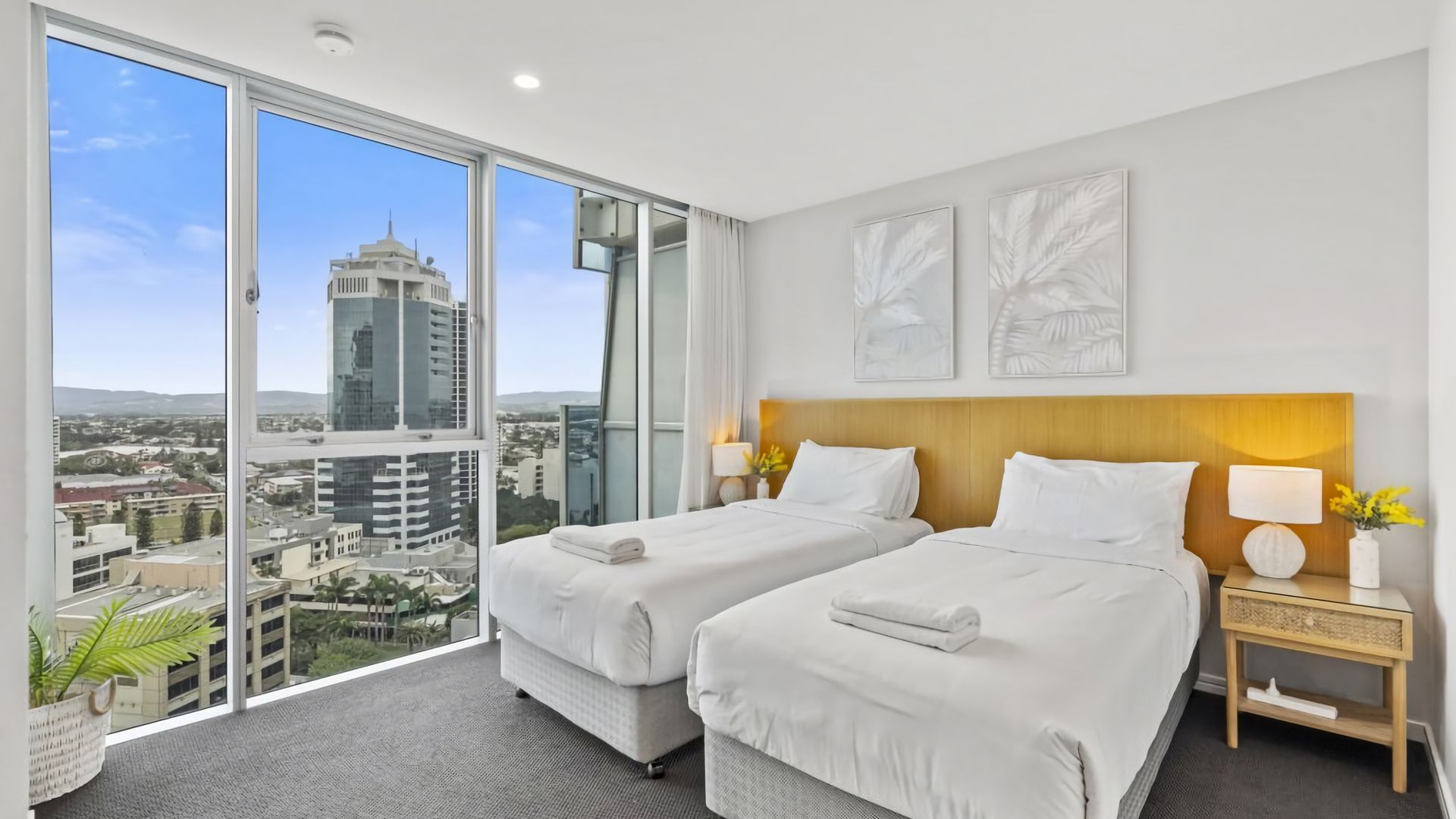 A hotel room with two beds, a large window offering a city view, and a nightstand with a lamp and flowers.
