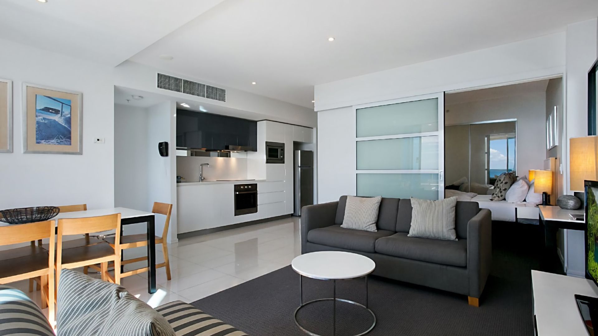 A modern living room with a grey sofa, a dining table with chairs, and a kitchen area with white cabinets and appliances.