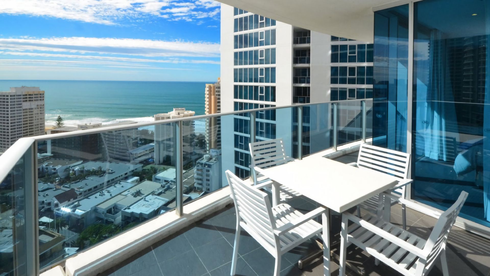 A balcony with a glass railing, white furniture, and a view of the ocean and cityscape.