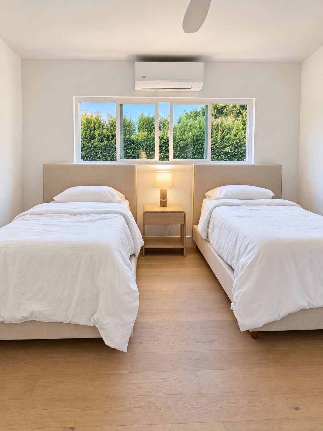A room with two beds, a nightstand, a lamp, and a window with a view of greenery.