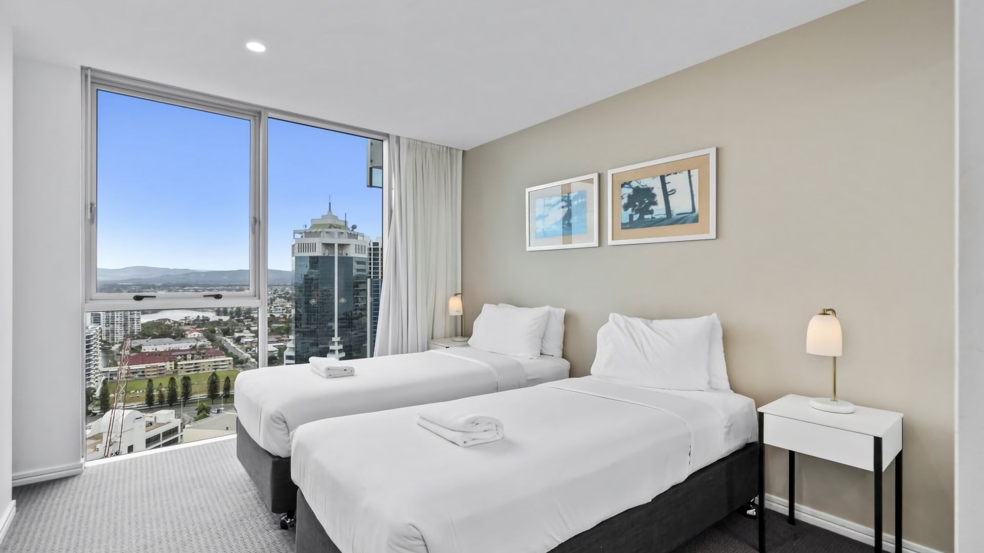 A hotel room with two beds, a large window with a city view, a bedside table with a lamp, and two framed pictures on the wall.