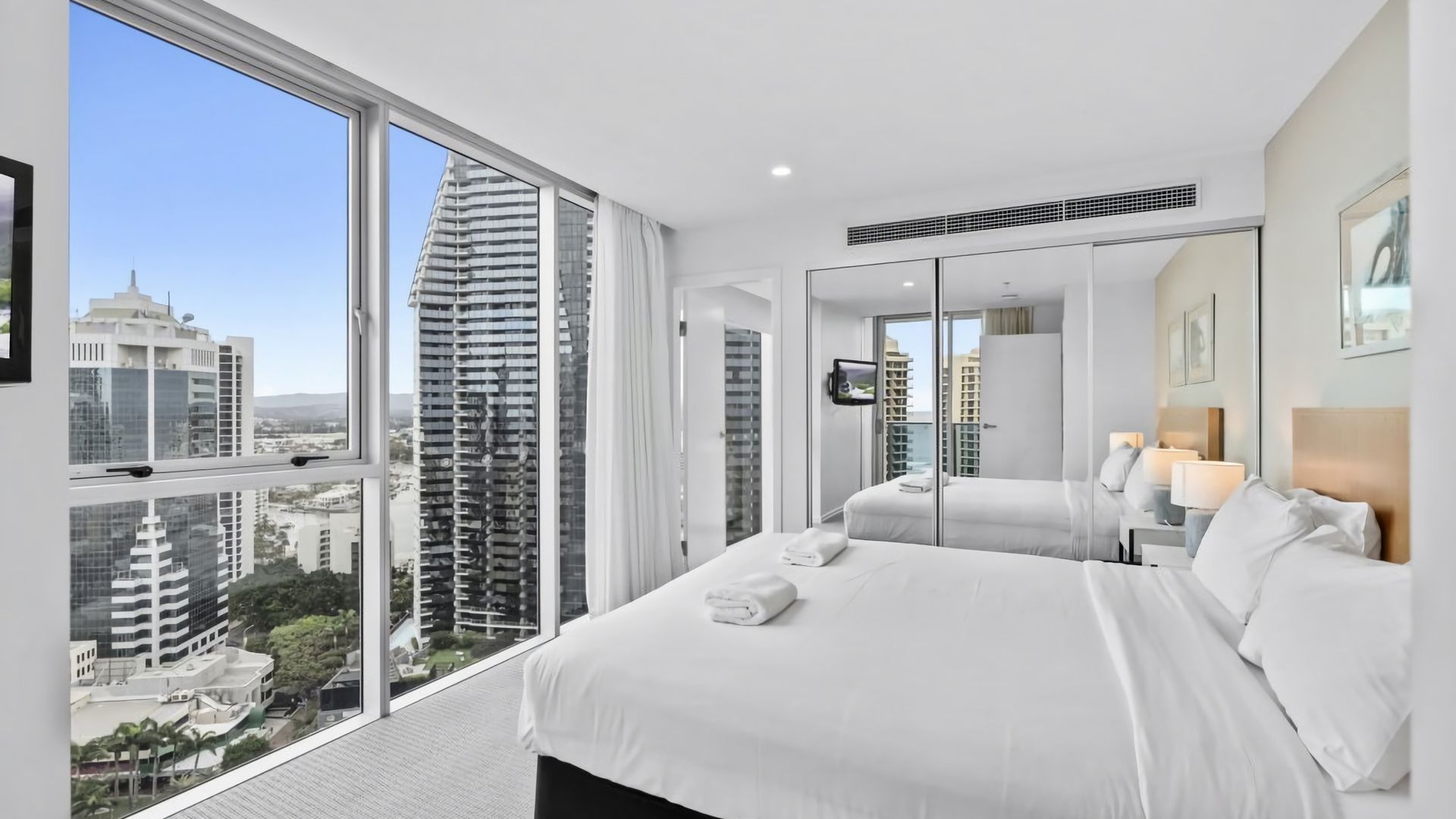 A hotel room with a large bed, a TV, a mirror, and a view of tall buildings.