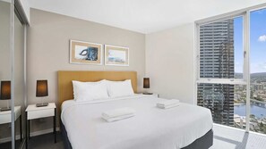 Ein Hotelzimmer mit einem großen Bett, zwei Nachttischen, einem Schminktisch und Blick auf eine Stadtlandschaft durch ein großes Fenster.