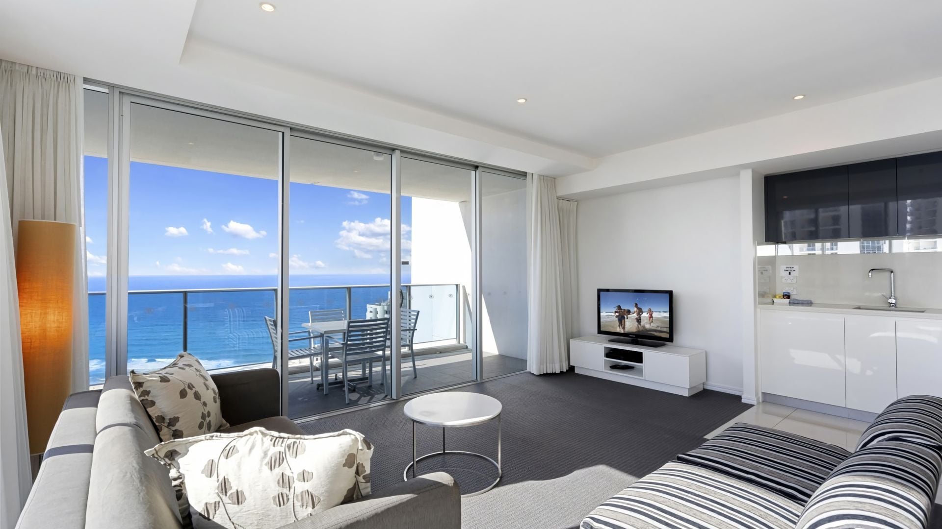 Una sala de estar moderna con balcón vistas al mar, un televisor y una zona de cocina.