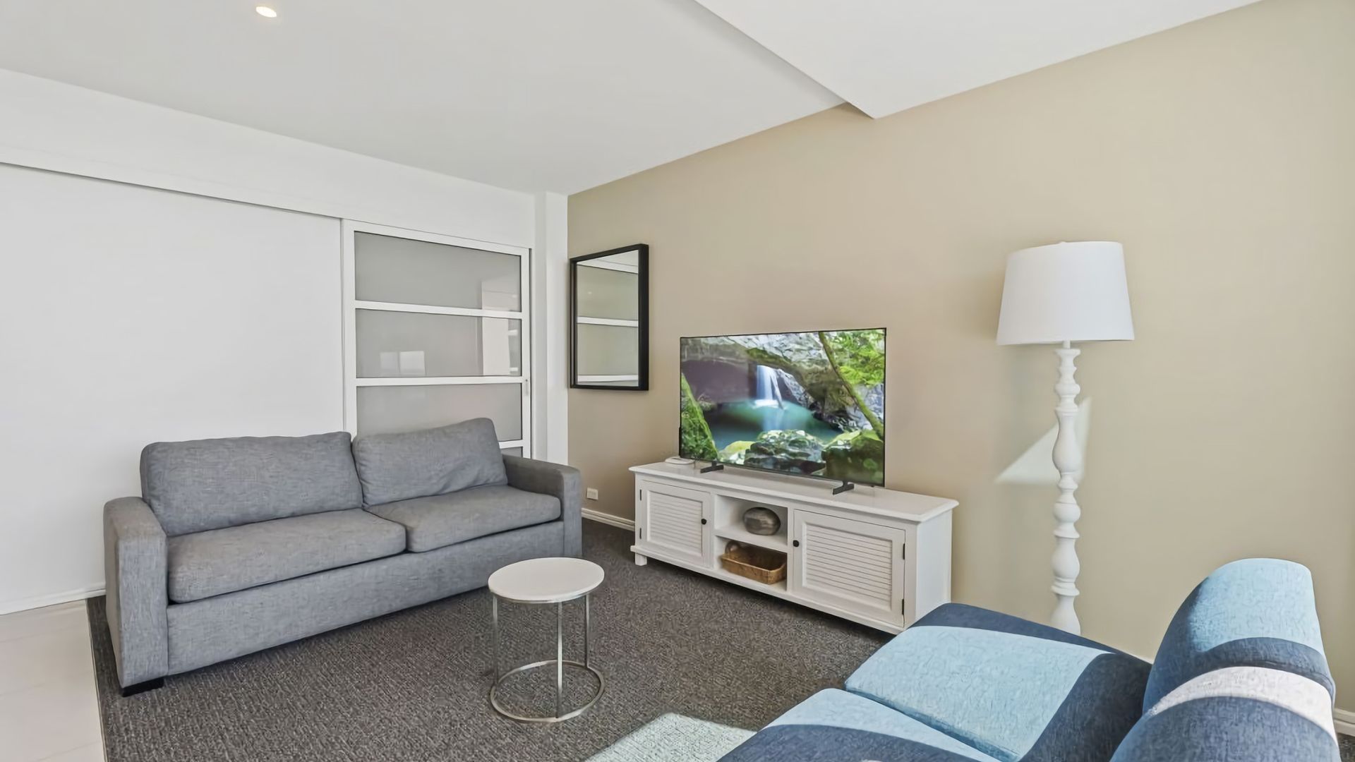 A modern living room with a grey sofa, a small round table, a TV on a white cabinet, and a lamp.